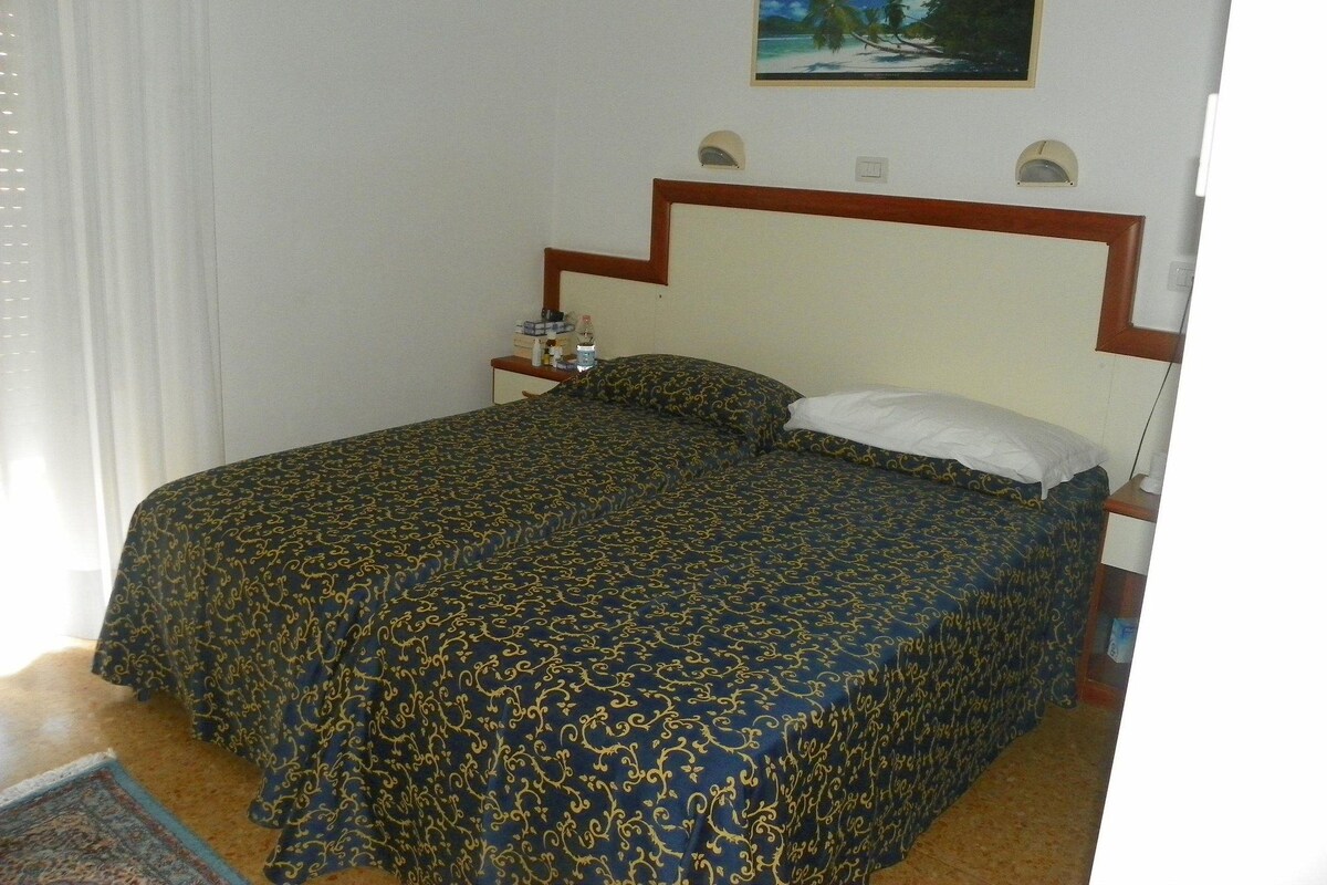Un lit bien fait, recouvert d’une couvre-lit à motifs, un tableau accroché au-dessus de la tête de lit, et une petite table de chevet avec une lampe et une bouteille.