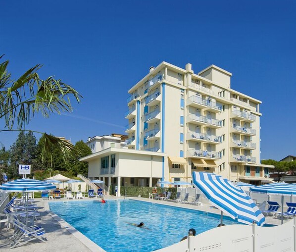 A hotel with a swimming pool, multiple balconies, and a clear blue sky.