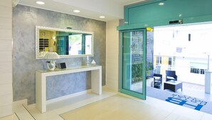 Un moderno vestíbulo de hotel con un mostrador de recepción, un espejo grande y una puerta de cristal que da a una zona de estar exterior.