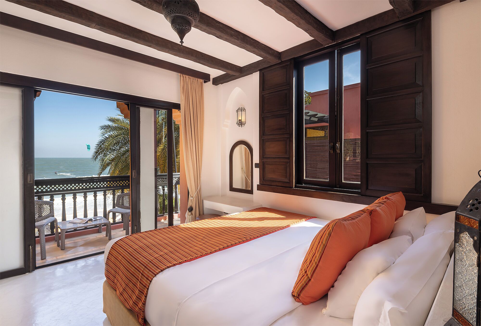 A hotel room with a bed, a balcony with a view of the sea, and a large window with dark wooden shutters.