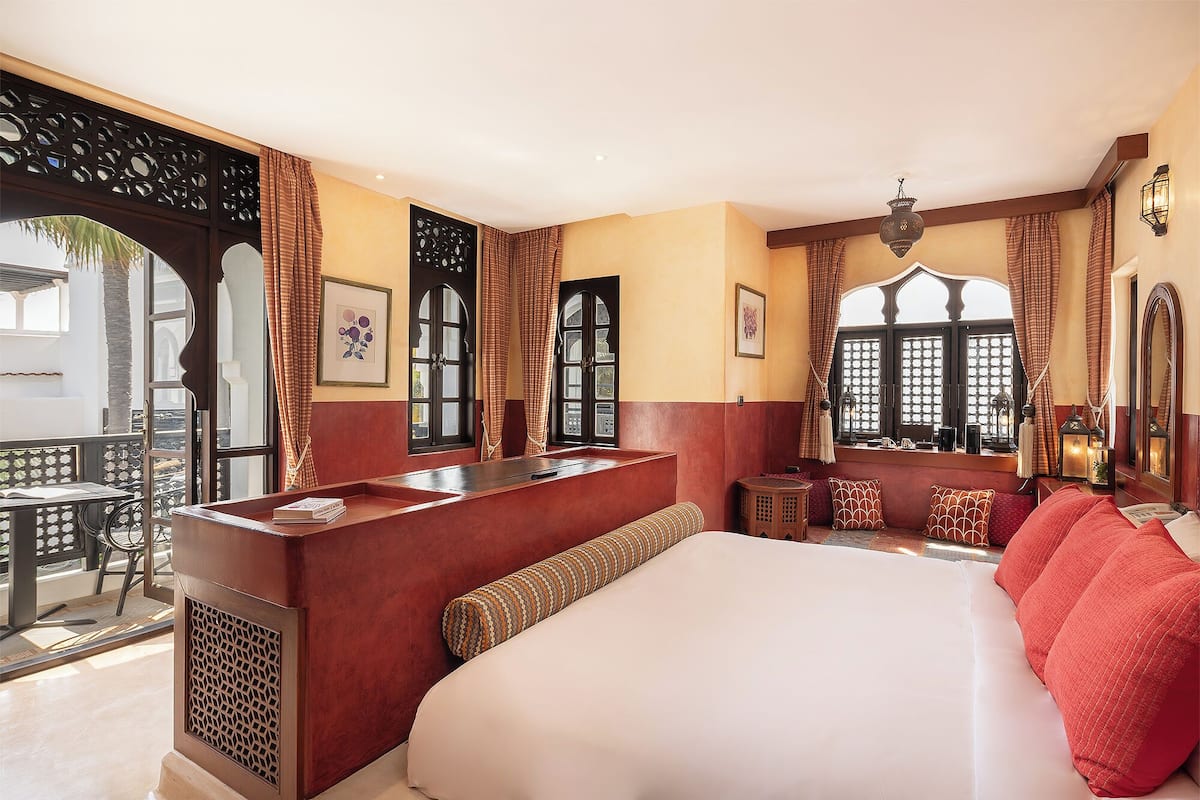 A hotel room with a large bed, a small seating area, a window with curtains, and a balcony with a view.