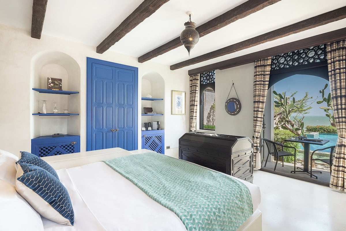 A bedroom with a bed, a blue door, a view of the sea, and a balcony with a table and chairs.