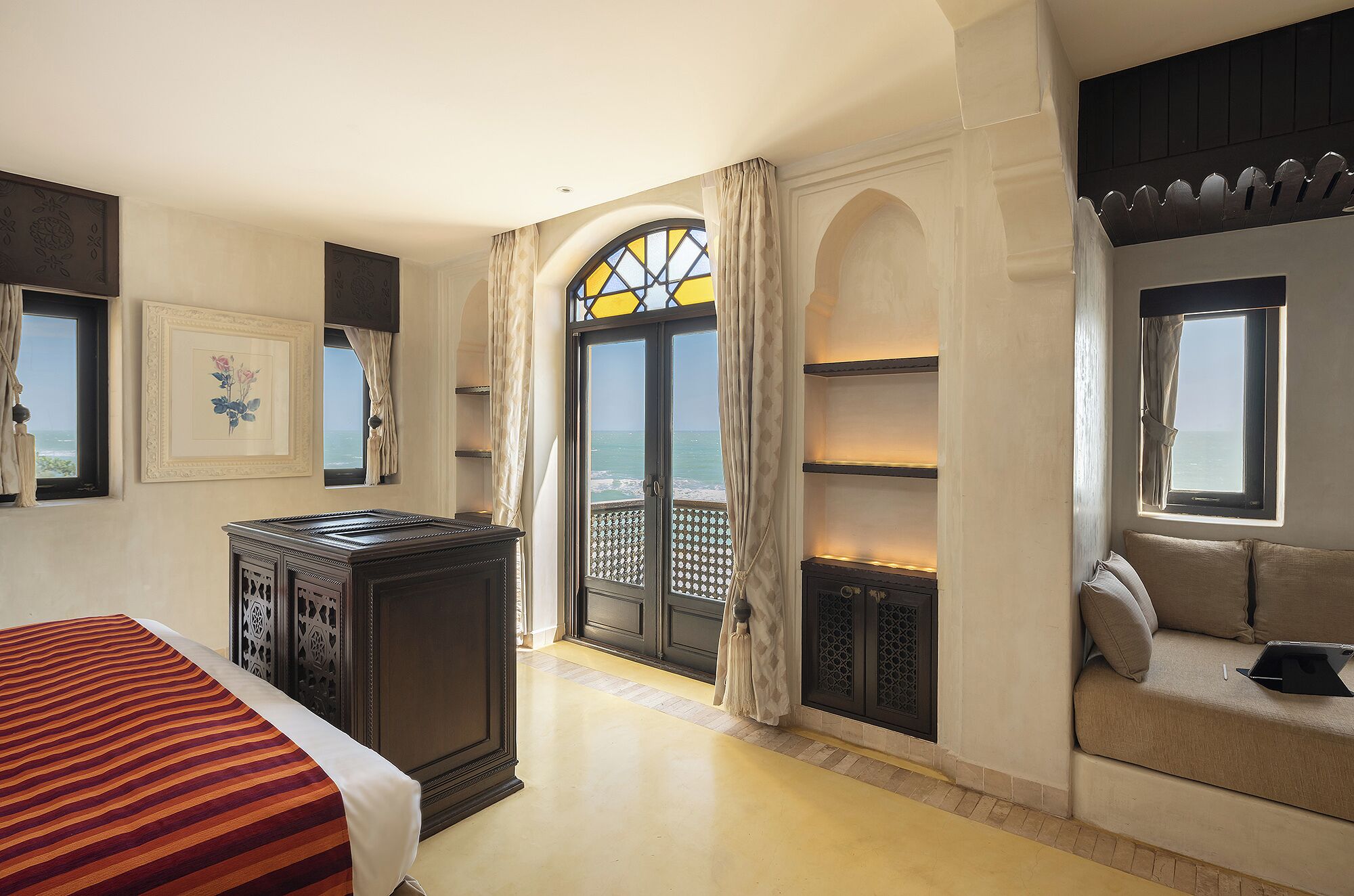 A hotel room with a bed, a sofa, a desk, and a view of the sea through large windows.