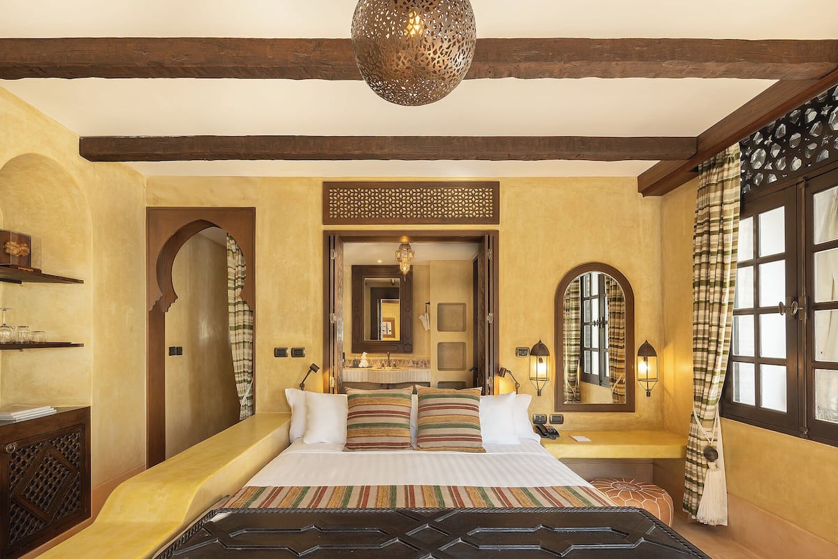 A bedroom with a large bed, wooden beams, a chandelier, and a view through a window with curtains.