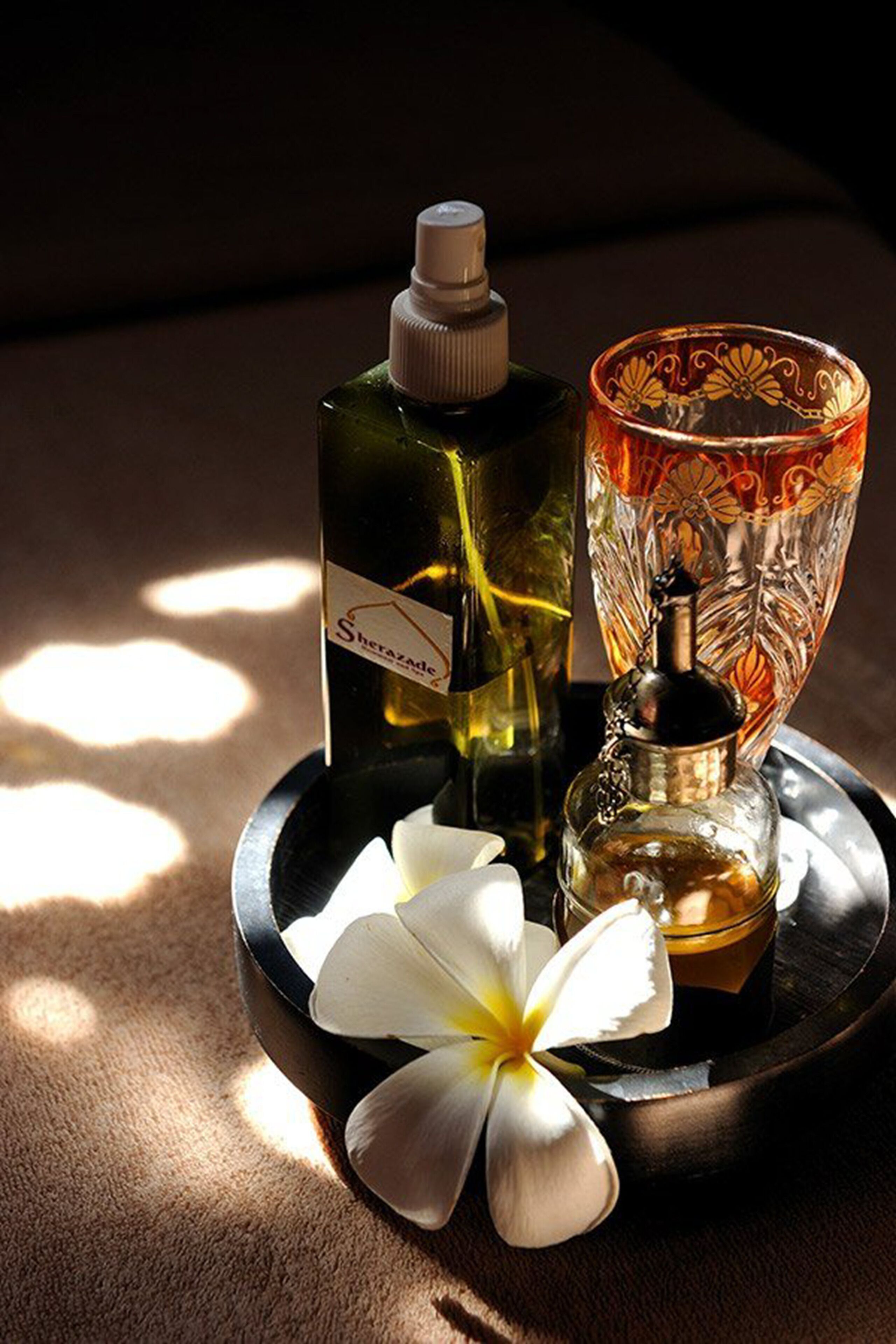 A tray with a bottle of Sherazade cologne, a glass with a decorative pattern, a small bottle, and a white flower.