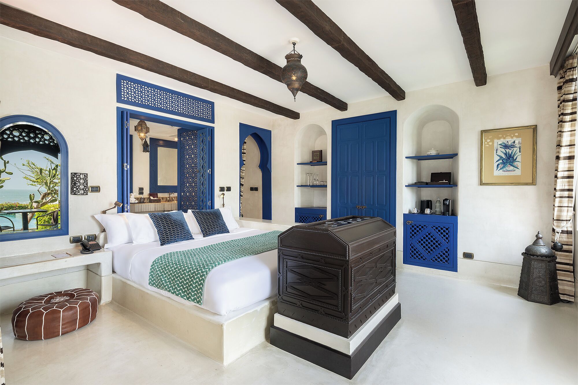 Een slaapkamer met een bed, nachtkastjes, een kaptafel en uitzicht op zee door een raam.