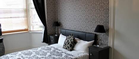 A neatly arranged bedroom with a patterned bedspread, two bedside tables with lamps, a window with curtains, and a patterned wallpaper.