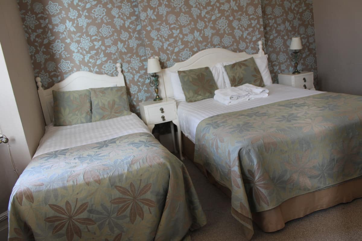A hotel room with two beds, floral-patterned bedding, a nightstand with a lamp, and a floral wallpaper.