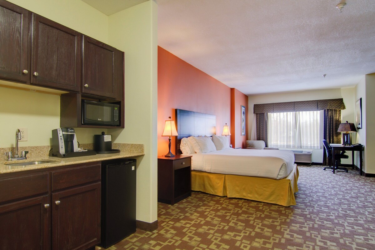 A hotel room with a bed, a desk, a chair, a desk lamp, a microwave, a coffee maker, and a sink.