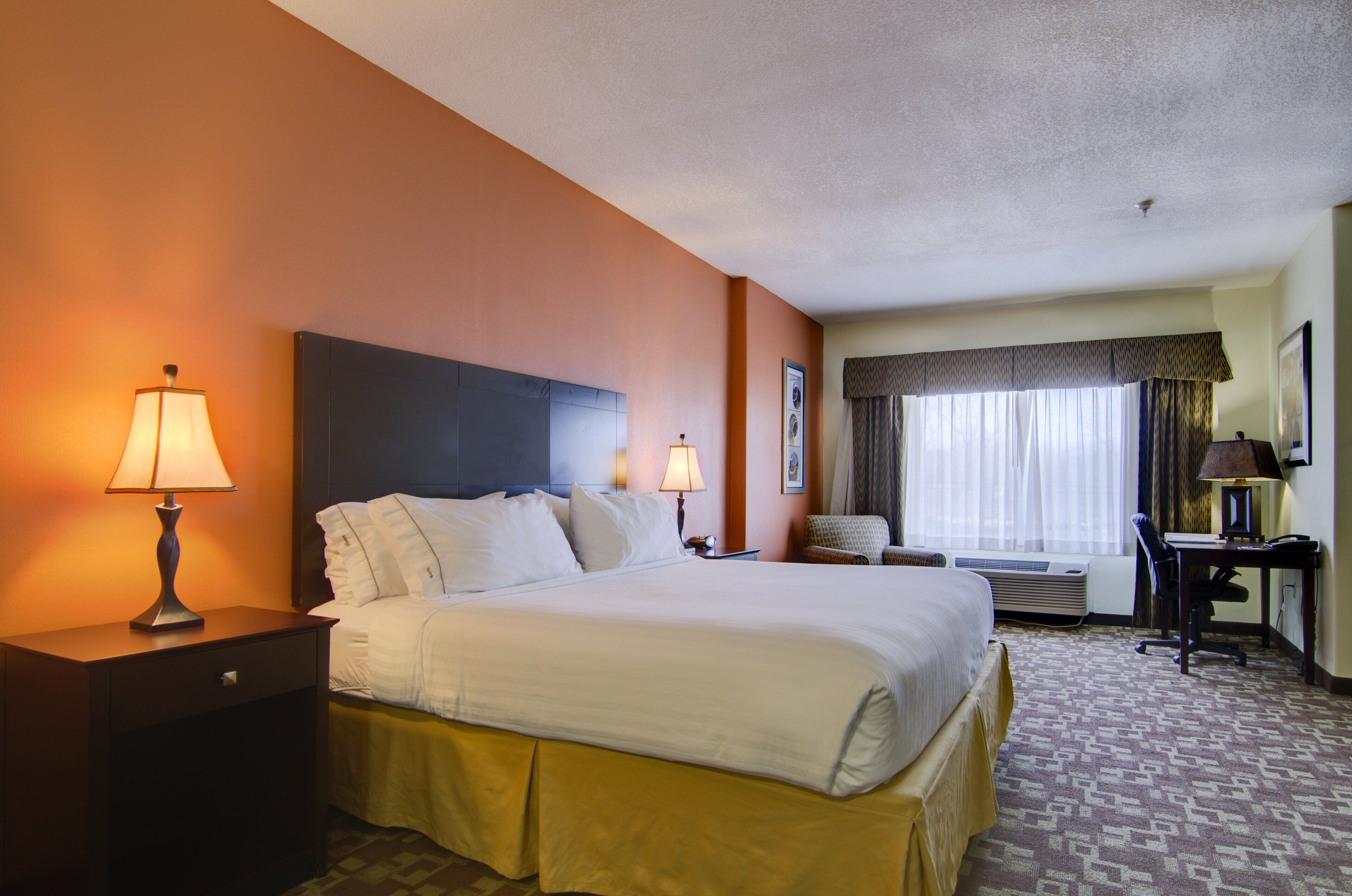 A hotel room with a large bed, two bedside lamps, a desk with a chair, and a window with curtains.