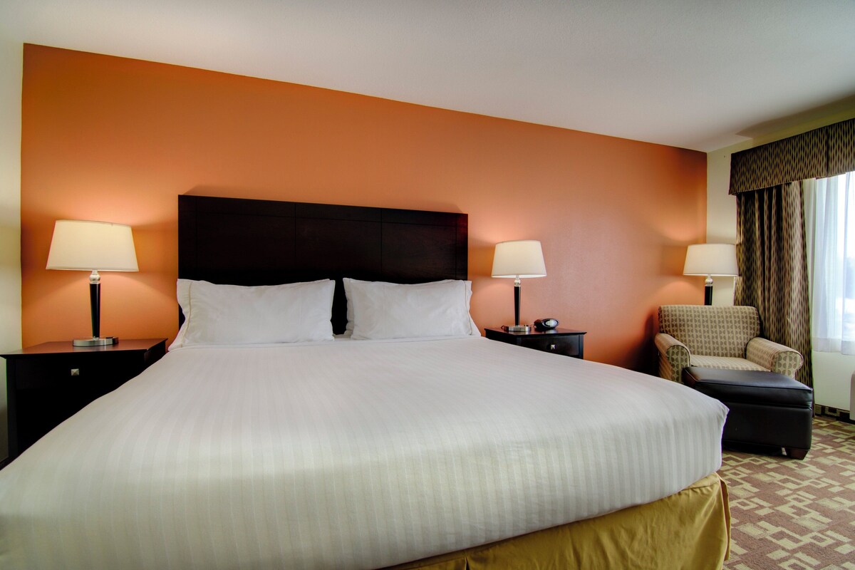 A hotel room with a large bed, two bedside tables with lamps, a chair, and a window with curtains.