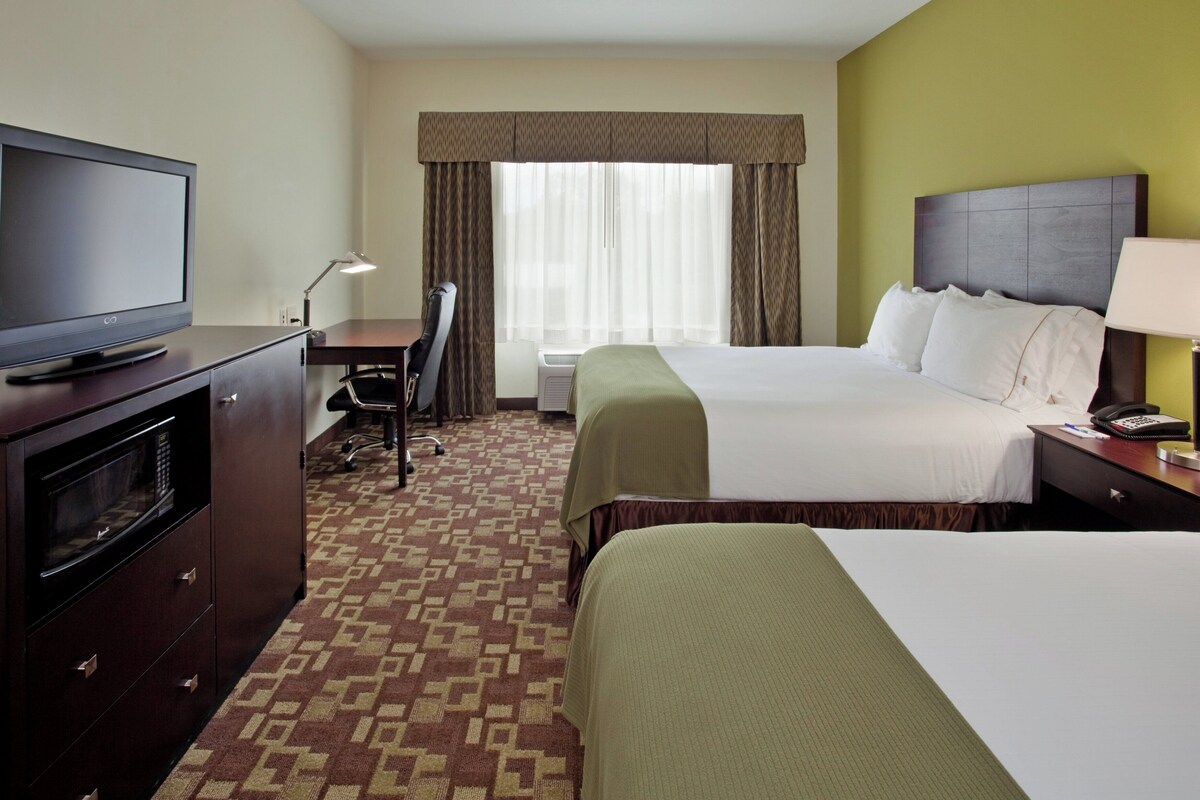 A hotel room with a television, desk, chair, two beds, and a window with curtains.