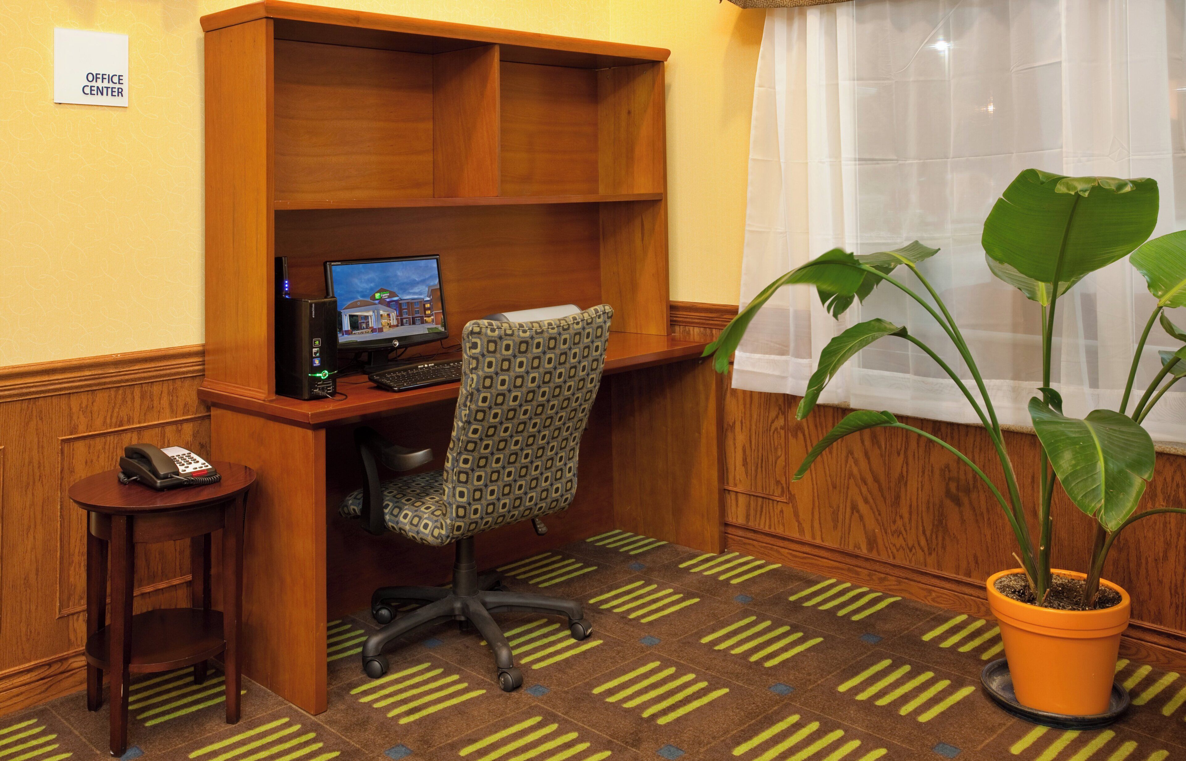 A hotel office center with a desk, computer, chair, telephone, and a potted plant.