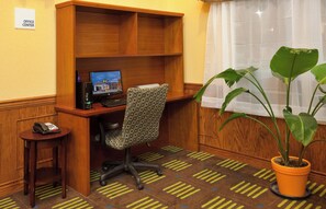 Espaço de escritório em hotel, com mesa, computador, cadeira, telefone e planta em vaso.