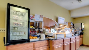 A breakfast buffet area with a refrigerated display of yogurt and milk, cereal options, and a variety of fruits.