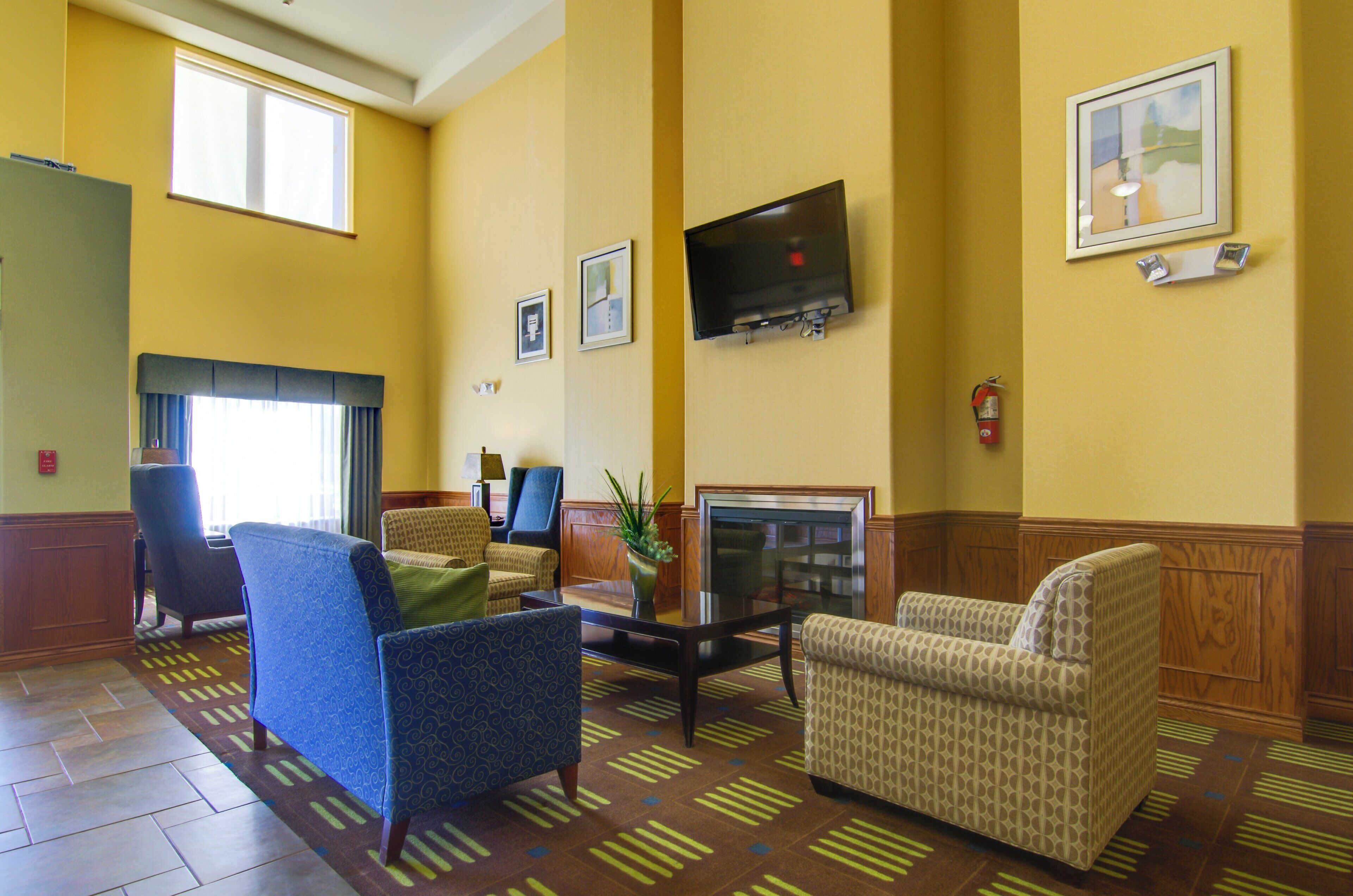 A hotel lobby with a flat-screen TV mounted on the wall, a fire extinguisher, and a window providing natural light.