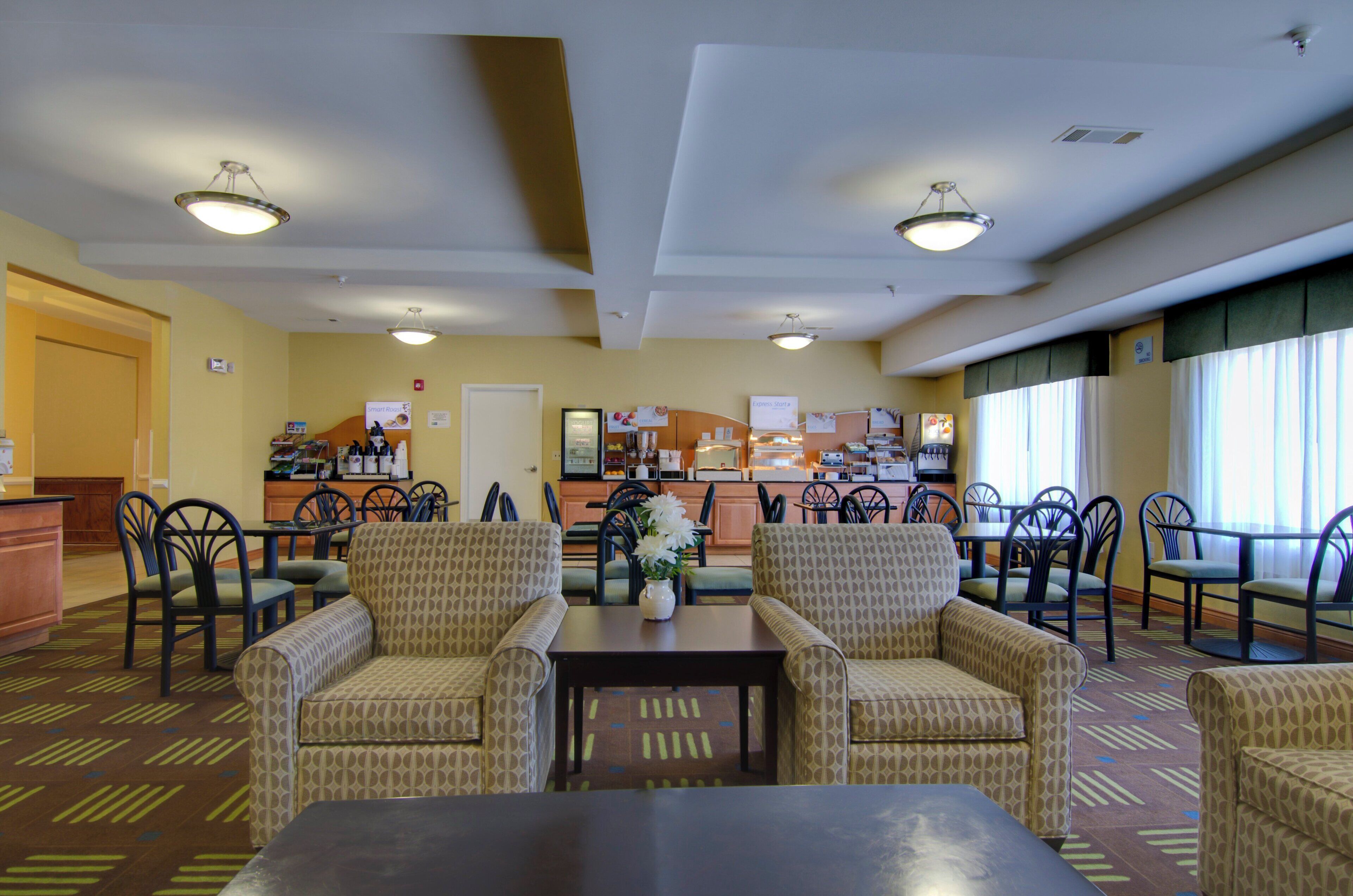 A hotel dining area with a central table, multiple chairs, a kitchen in the background, and a window with curtains.