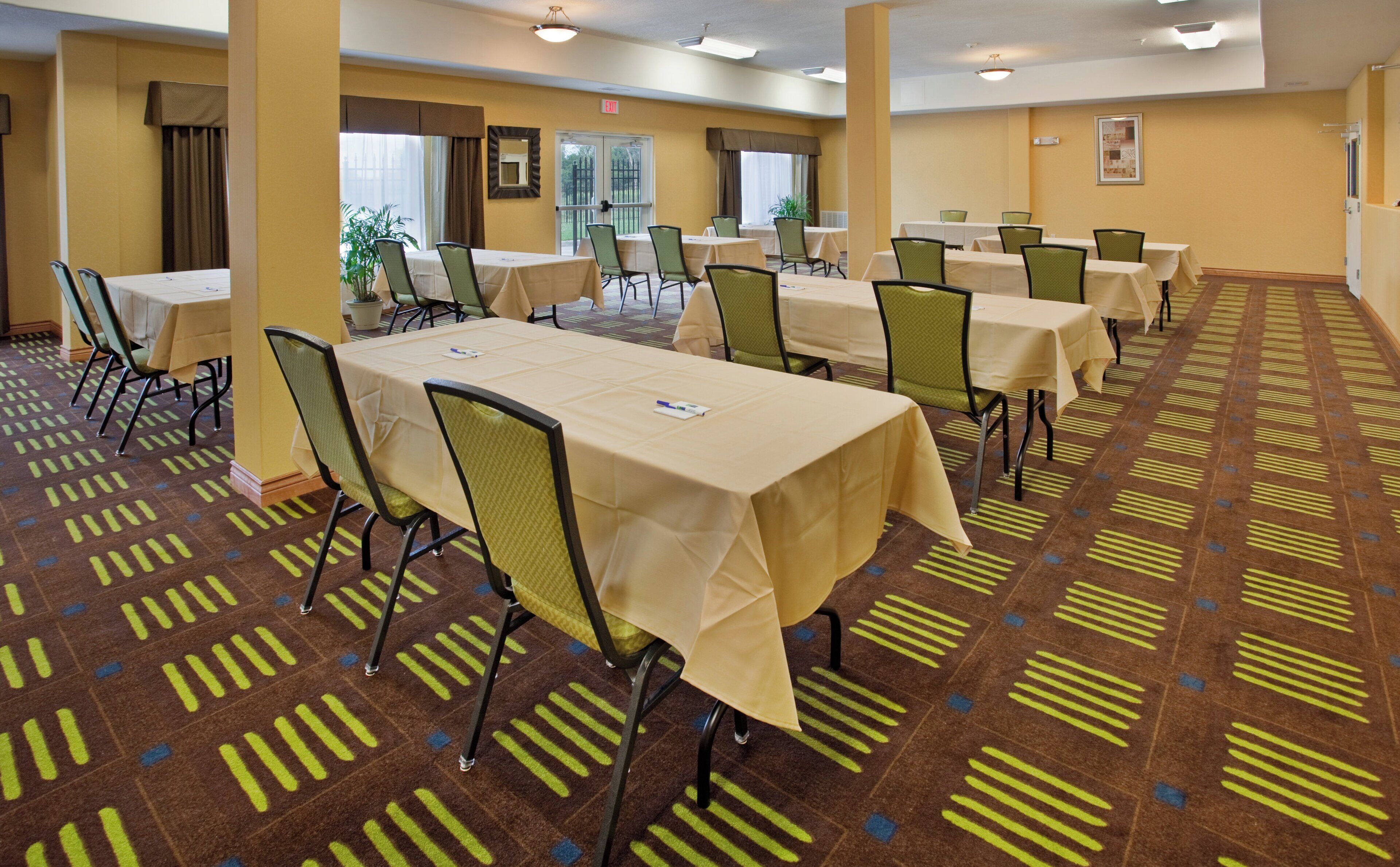 A hotel conference room with multiple tables and chairs, carpeted floor, and windows with curtains.