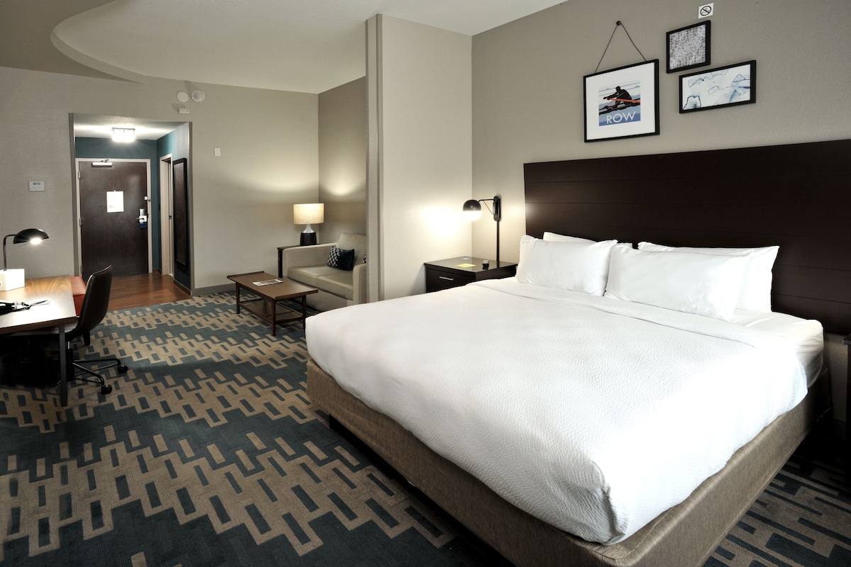A hotel room with a large bed, a desk with a chair, a nightstand, and a lamp. There are framed pictures on the wall.