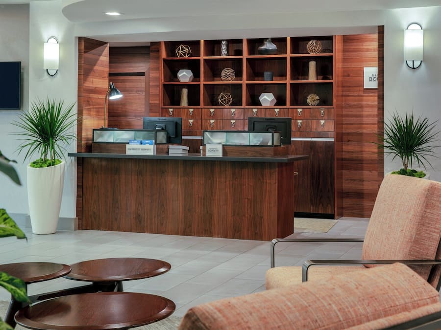 Una zona de recepción con un mostrador de madera, un televisor de pantalla plana y un letrero que dice 'MARRIOTT BONVIVI'. Hay dos plantas en macetas, un área de descanso con mesas y sillas redondas, y un estante de pared con objetos decorativos.
