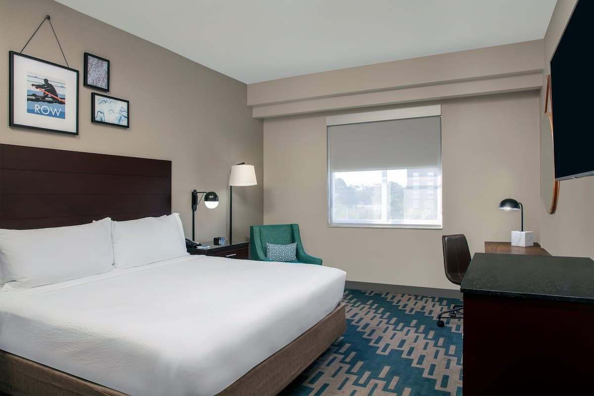 A hotel room with a large bed, a desk, a chair, a window, and framed pictures on the wall.