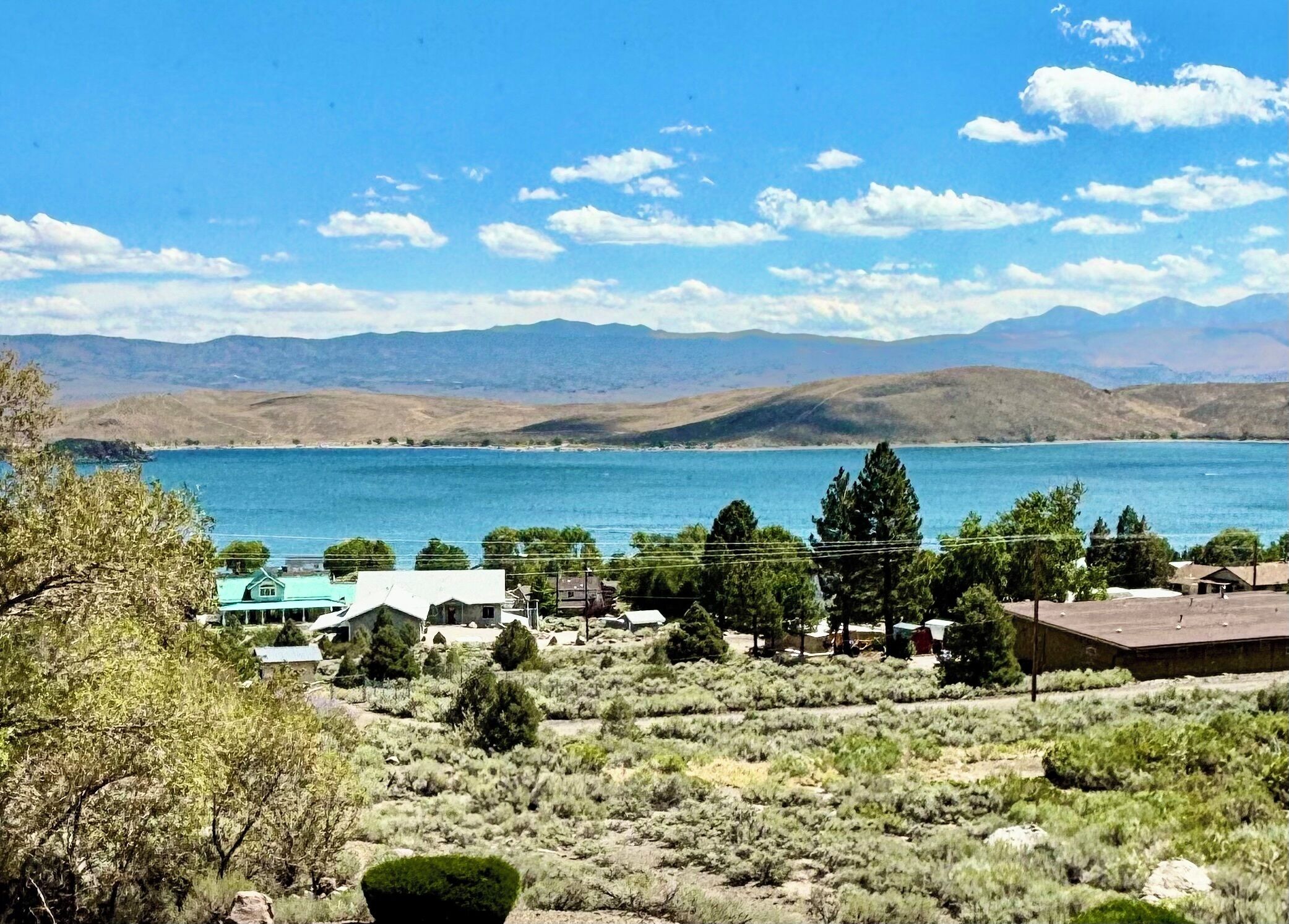 A lakeside property with a view of mountains, surrounded by sparse vegetation and a few buildings.