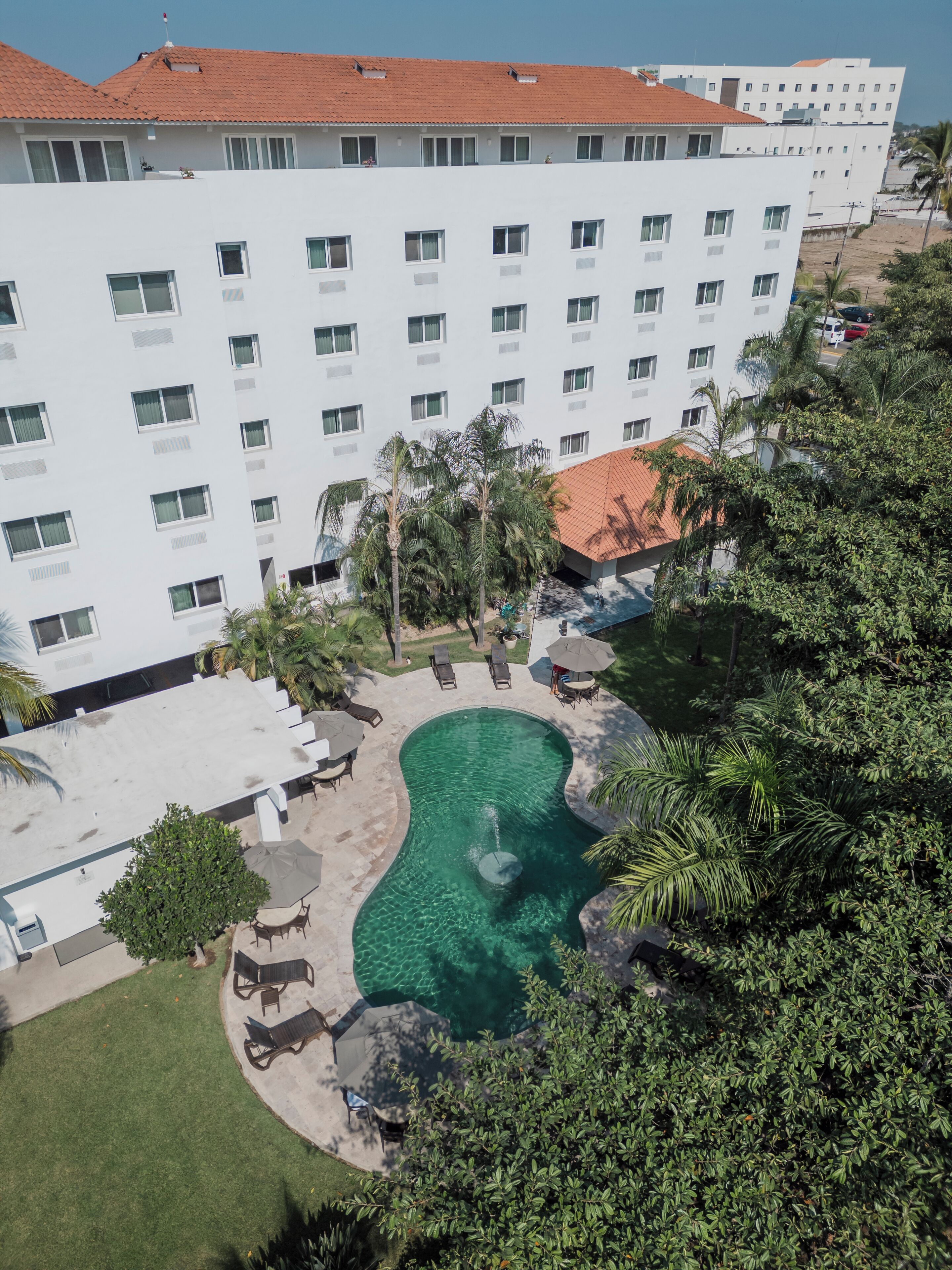 A hotel with a swimming pool surrounded by lounge chairs and umbrellas, a well-maintained lawn, and a red-tiled roof.