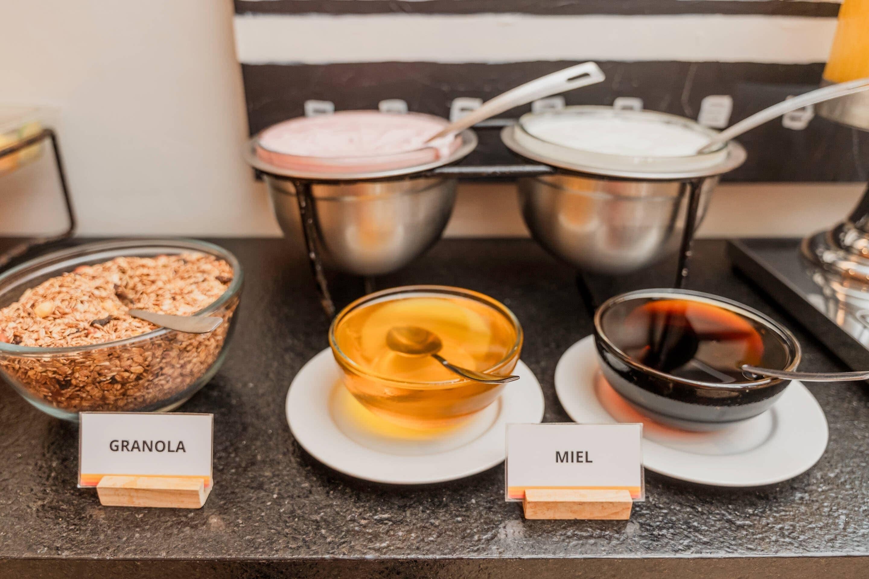 Un bufé de desayuno con recipientes identificados con granola, muesli y un líquido oscuro, posiblemente café o té.