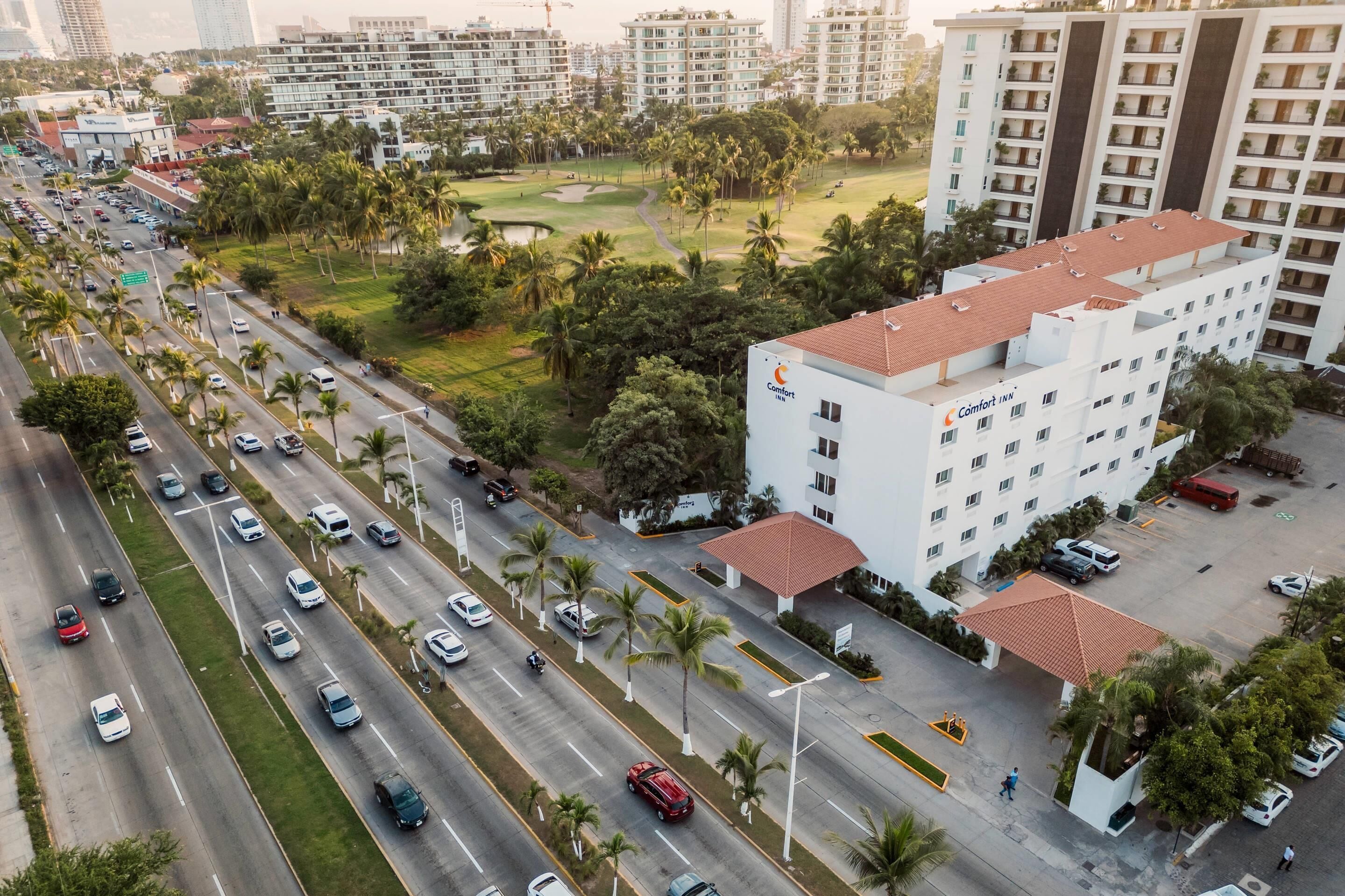 Un hotel de varios pisos con techo plano, rodeado por una carretera con coches y palmeras.