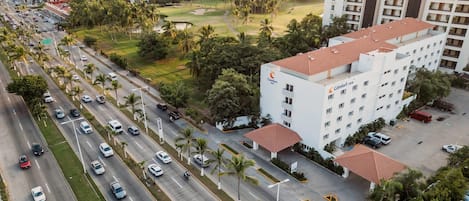 A multi-story hotel with a flat roof, surrounded by a road with cars and palm trees.