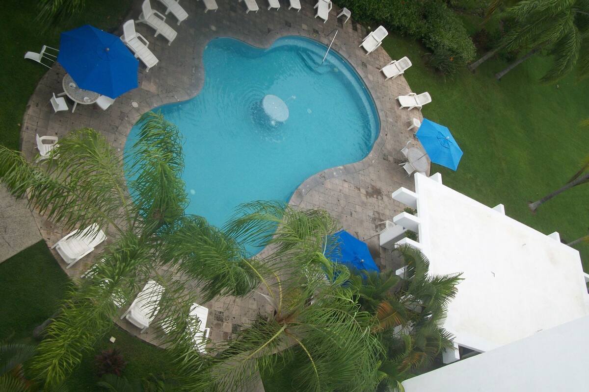 Eine Vogelperspektive zeigt einen Swimmingpool, umgeben von weißen Liegestühlen und blauen Sonnenschirmen, mit Palmen und einem weißen Gebäude im Hintergrund.