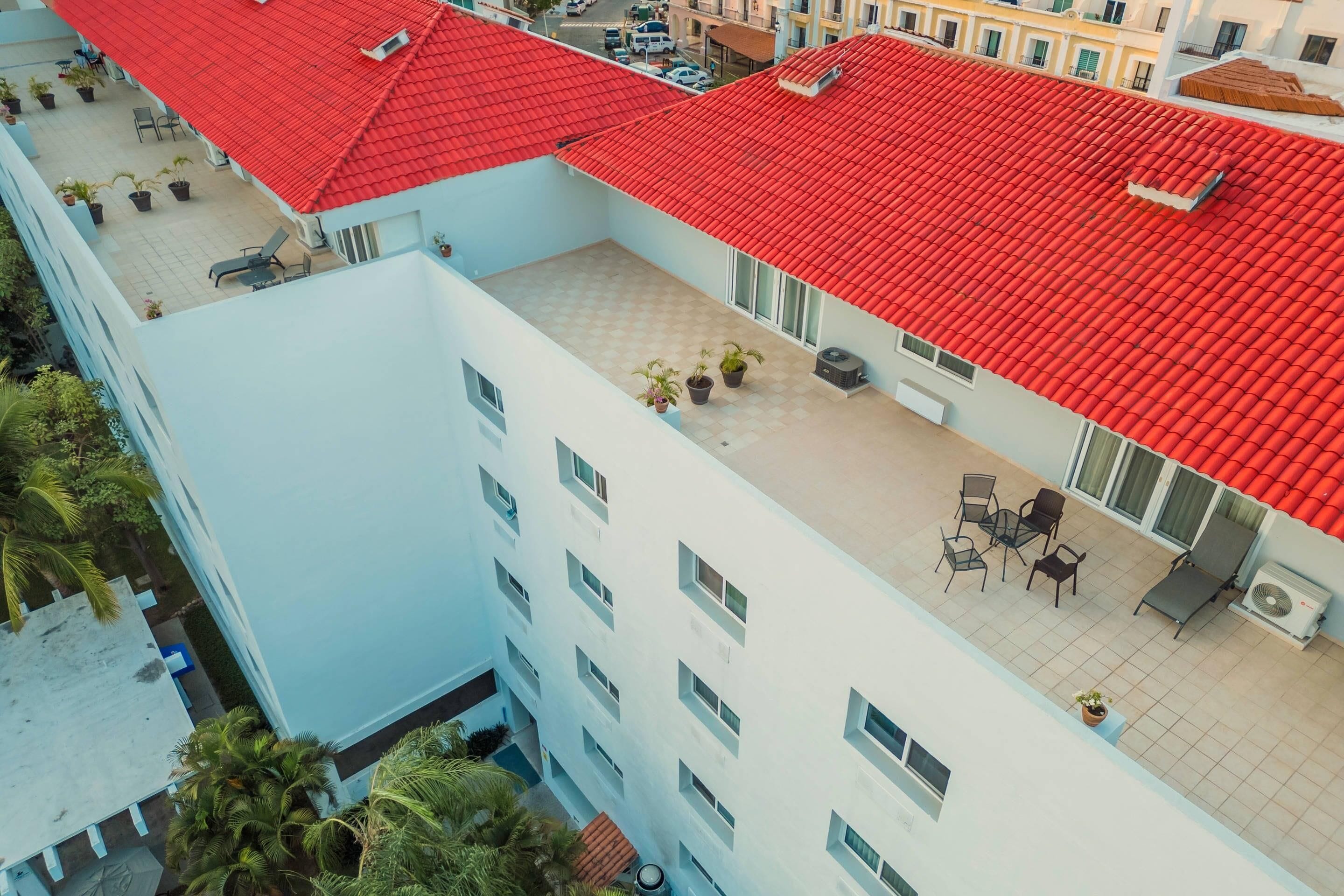 Um edifício de vários andares com telhado de telhas vermelhas, área externa com mesas e cadeiras, e plantas em vasos.