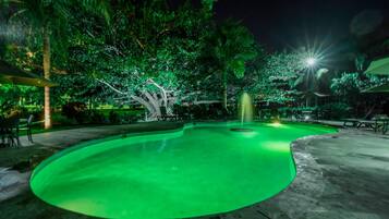 Une piscine nocturne illuminée de lumières vertes, entourée d’arbres et dotée d’un espace de détente extérieur.