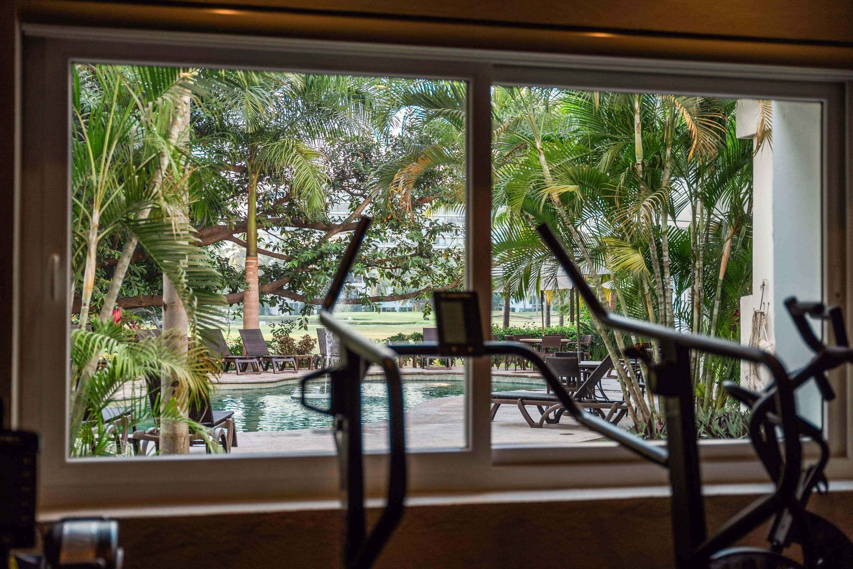 A view of a pool area with lounge chairs and a water feature, framed by a window with exercise equipment in the foreground.