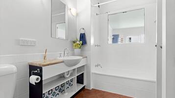 A modern bathroom with a white sink, mirror, and bathtub. There is a towel rack with a blue towel, and a small shelf with decorative items.