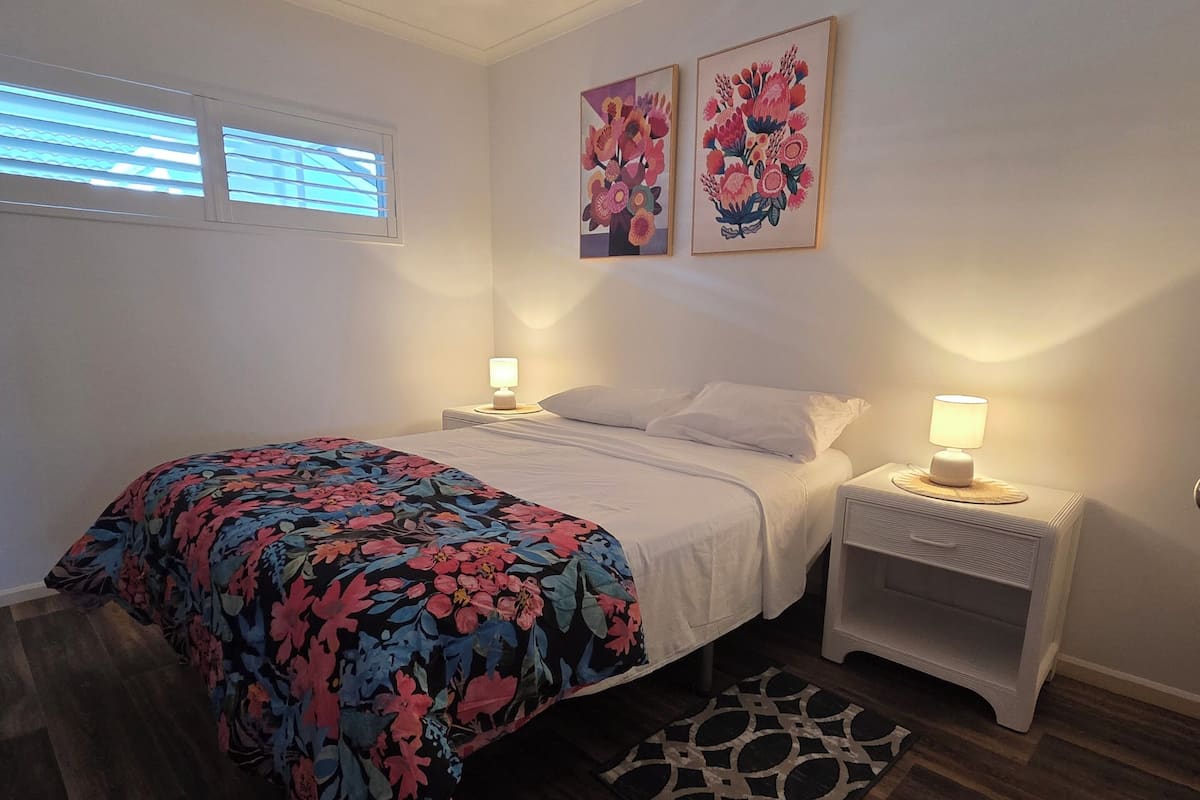 Une chambre à coucher comprenant un lit, deux tables de chevet, un ventilateur de plafond et deux tableaux accrochés au mur.