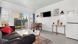 Una sala de estar moderna con un sofá, una mesa de centro y un comedor con una mesa y sillas. Hay un televisor montado en la pared, un refrigerador y una ventana con persianas.