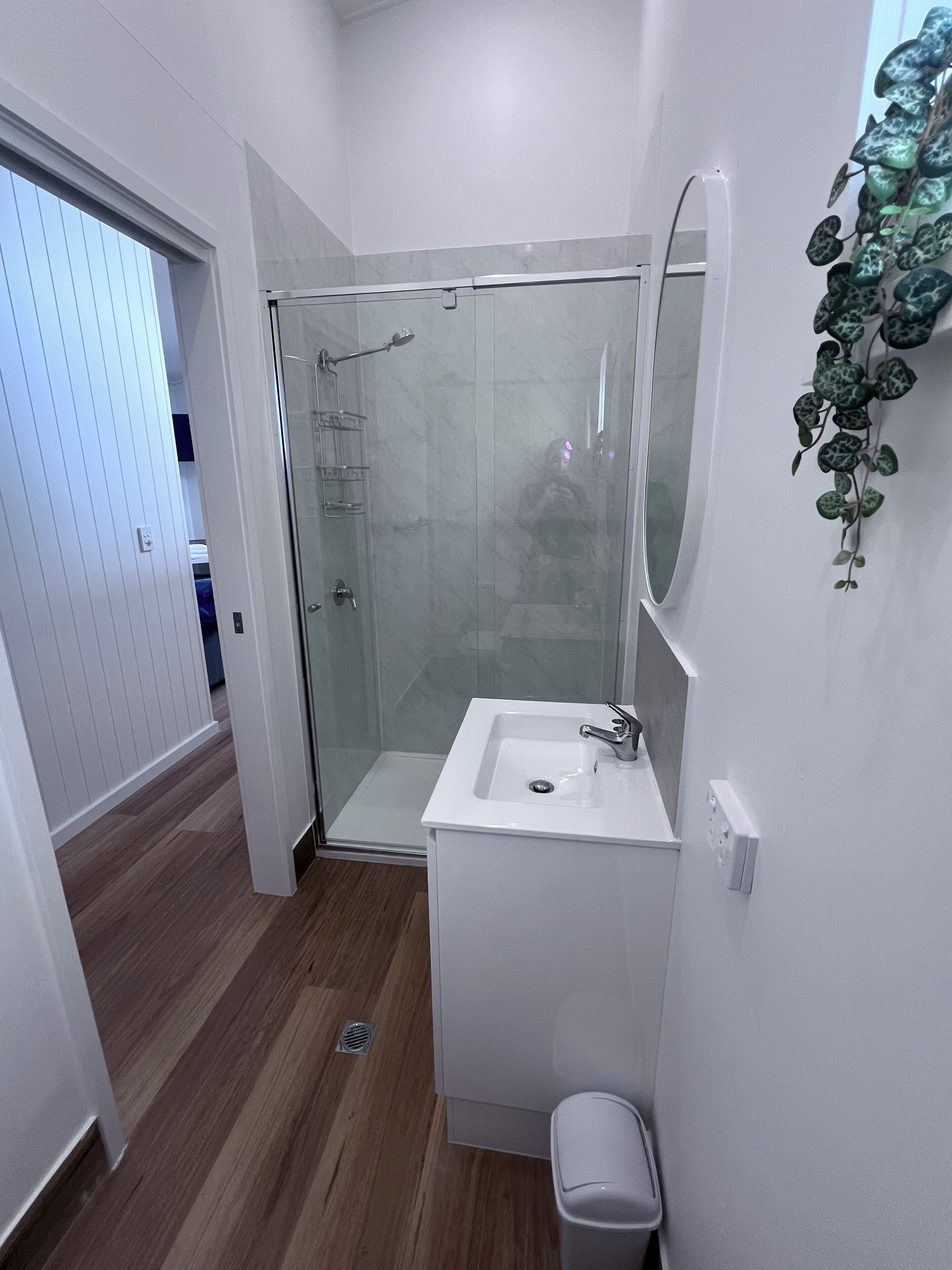Ein modernes Badezimmer mit Duschabtrennung aus Glas, einem weißen Waschbecken und einer kleinen Mülleimer.