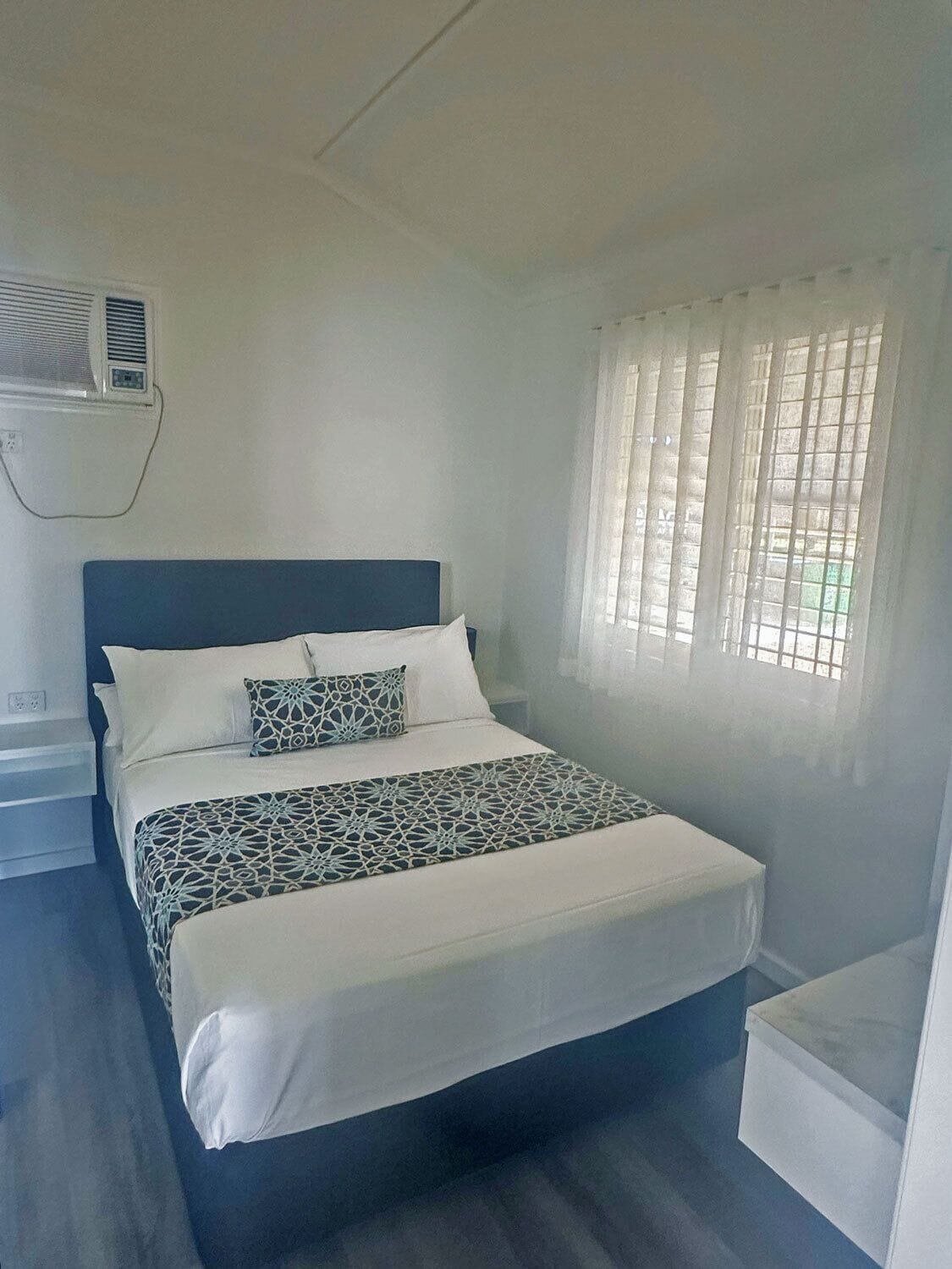 Habitación individual con una cama y cabecera azul oscuro, ropa de cama blanca, una manta estampada y una ventana con persianas verticales.