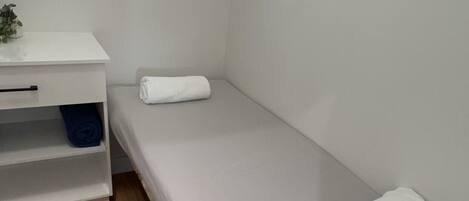 A small, single-bed room with a white bed, a desk with a shelf, and a window.