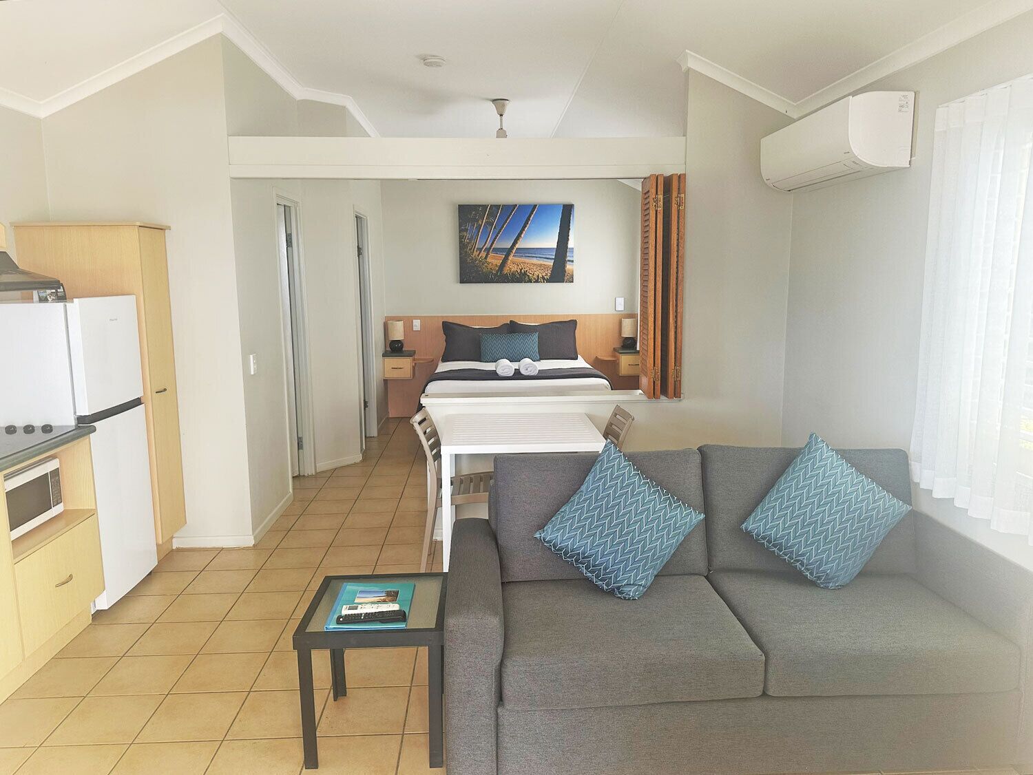 A compact hotel room with a kitchenette, a bed with a headboard, a gray sofa with patterned cushions, and a painting on the wall.