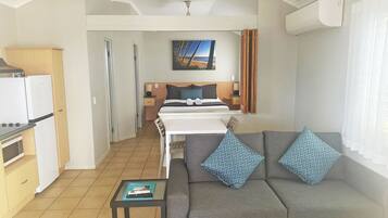 A compact hotel room with a kitchenette, a bed with a headboard, a gray sofa with patterned cushions, and a painting on the wall.