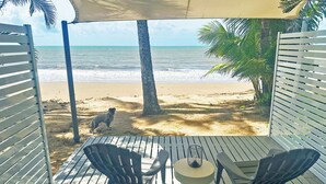 Une terrasse avec deux chaises et une table, donnant sur une plage où l’on aperçoit un chien et des palmiers.