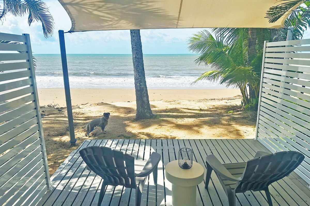 Une terrasse avec deux chaises et une table, donnant sur une plage où l’on aperçoit un chien et des palmiers.