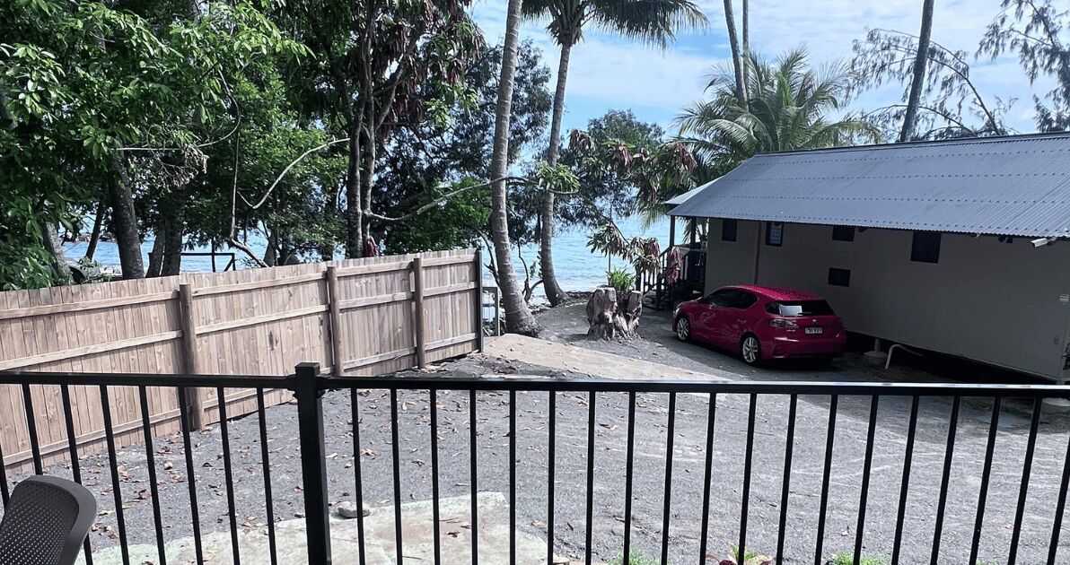 A view of a beachside property with a wooden fence, a red car, and a building with a metal roof.