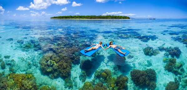 Two people floating on blue mats in clear blue water with coral reefs, a small island in the background, and a clear sky. - Green Island Resort (Green Island)
