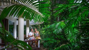 Una pareja sentada en una terraza de madera con vistas a abundante vegetación y una casa al fondo.