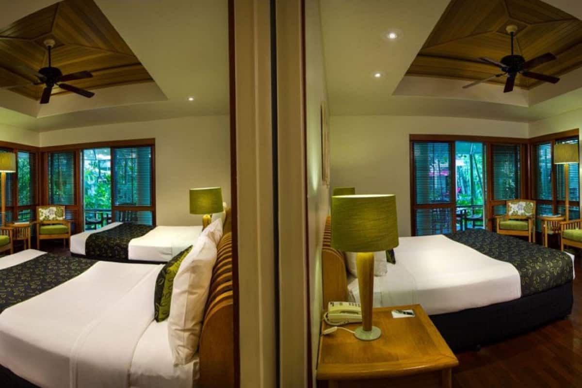 Habitación de hotel con espejos, dos camas, piso de madera, ventiladores de techo y ventanas grandes con persianas.
