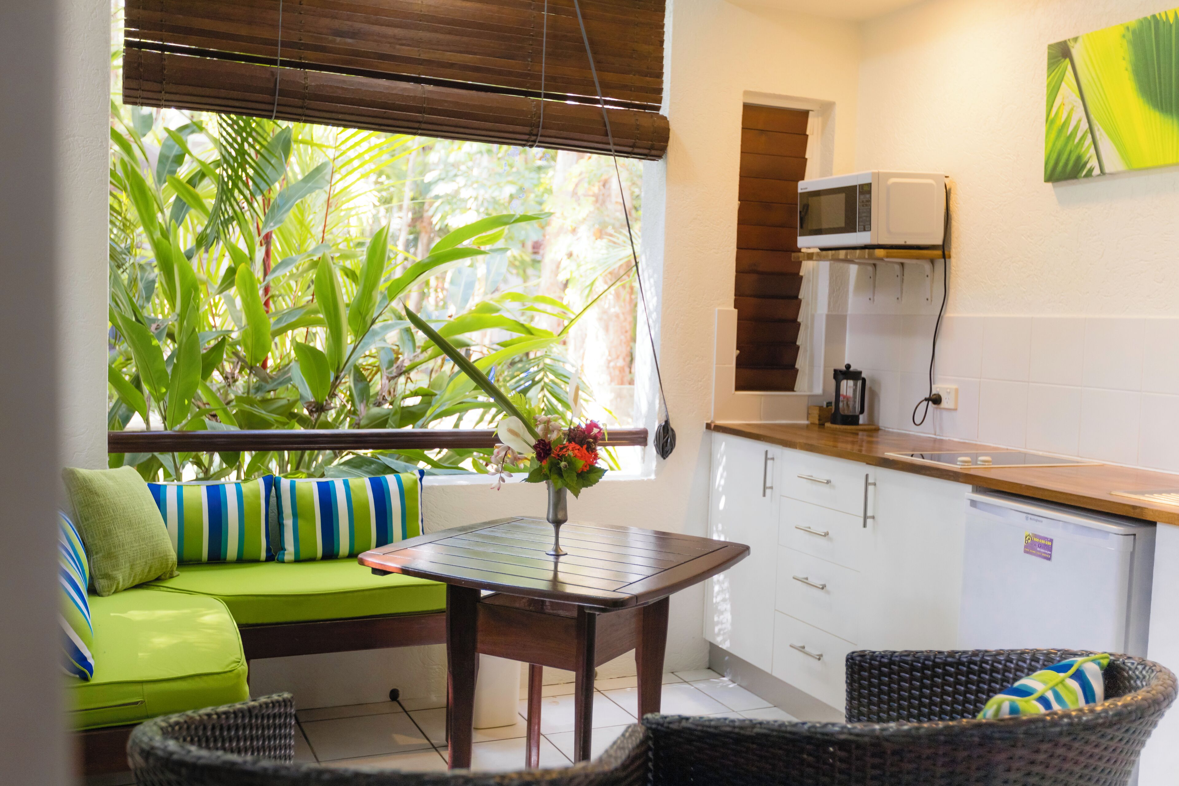Una cocina compacta con armarios blancos, un microondas y un refrigerador pequeño. Una mesa de comedor con un florero está ubicada cerca de una ventana con vista a follaje verde.