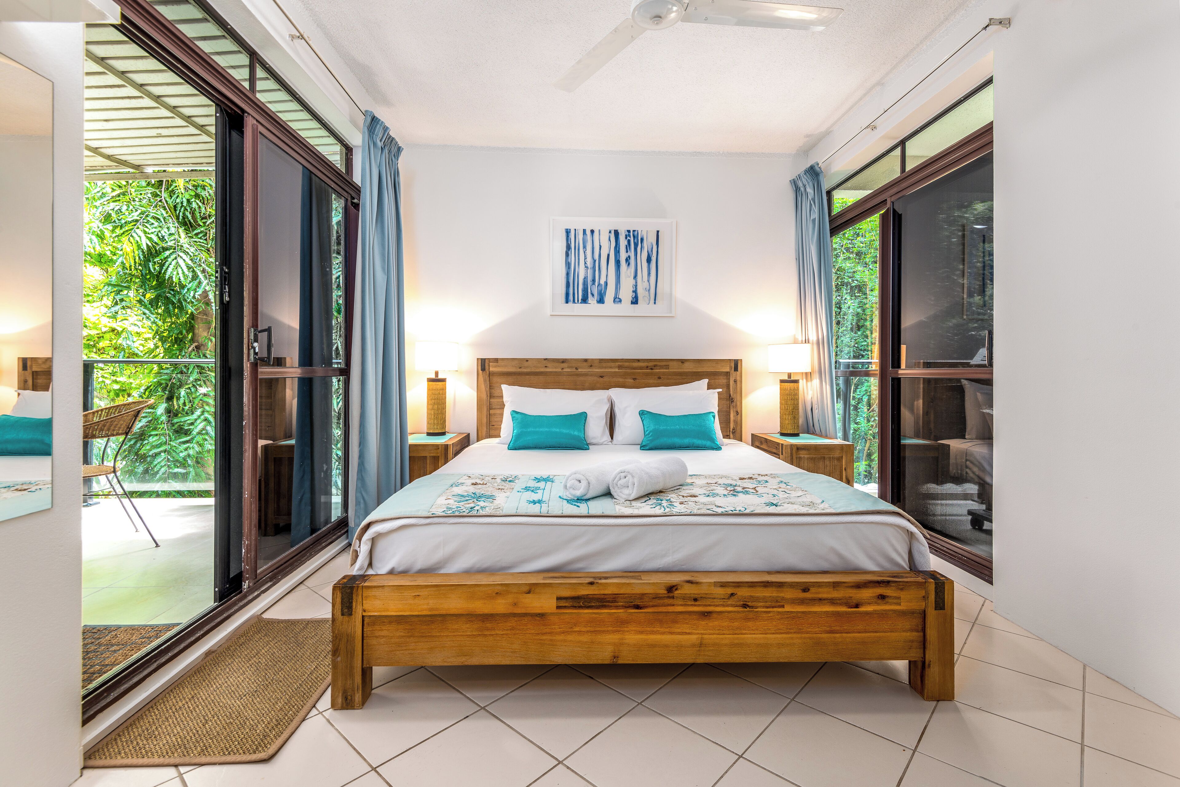 A bedroom with a wooden bed, a view of greenery through a sliding door, and a ceiling fan.